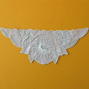 May include: A white lace trim with a floral design. The trim features three flowers, two smaller flowers on the sides and a larger flower in the center. The trim is perfect for adding a touch of elegance to any project.