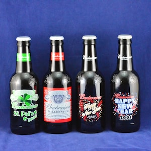 May include: Four Budweiser beer bottles with white caps. The bottles have labels for St. Patrick's Day 2000, Millennium, New Year 2002, and Happy New Year 2001. The bottles are black with colorful labels.