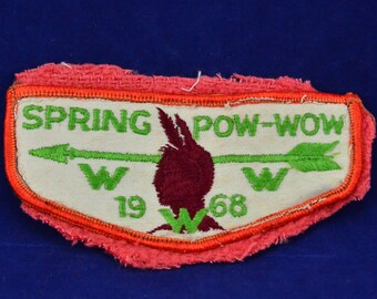 May include: A white fabric patch with green embroidered text that reads "SPRING POW-WOW" and a green arrow with a red bird in the center. The text "19 W 68" is also embroidered in green below the bird. The patch has a red border.