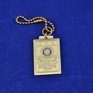 Könnte beinhalten: Ein rechteckiger, goldfarbener Schlüsselanhänger mit der Aufschrift "THEIR GLORY SHALL NEVER FADE". Der Anhänger zeigt das Emblem der American Legion in Blau und Weiß mit drei Sternen darüber. Eine goldene Kugelkette ist befestigt.