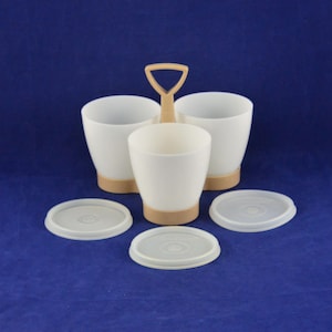 May include: A white plastic egg cup set with a tan handle and base. The set includes three egg cups and three clear lids. The egg cups are arranged in a carrier with a handle. The background is a solid blue.