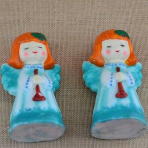 May include: Two small ceramic angel figurines with red hair, blue dresses, and white wings. Each angel is holding a red trumpet.