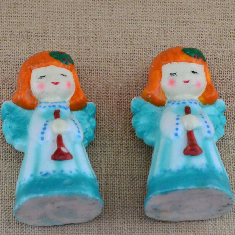 May include: Two small ceramic angel figurines with red hair, blue dresses, and white wings. Each angel is holding a red trumpet.