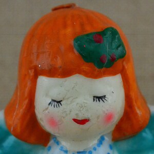 May include: A ceramic figurine of a girl with orange hair, closed eyes, and a green and red bow. She is wearing a blue and white dress.