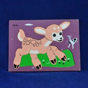 Può includere: Un puzzle in legno raffigurante un cerbiatto rosa chiaro con zoccoli neri e una farfalla a pois bianchi. I pezzi del puzzle sono ambientati su uno sfondo viola con una collina verde. La parola "Sifo" è stampata nell'angolo in alto a sinistra.