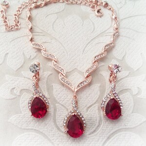 Puede incluir: Un conjunto de collar y pendientes de oro rosa con una piedra roja en forma de lágrima rodeada de brillantes transparentes.