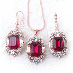 May include: A rose gold necklace and earrings set with a large, red, emerald-cut gemstone surrounded by smaller clear rhinestones.