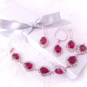 Könnte beinhalten: Ein Schmuckset aus roségold mit einem Armband, einer Halskette und Ohrringen. Jedes Stück hat einen großen, tropfenförmigen roten Strassstein, der von kleineren klaren Strasssteinen umgeben ist.
