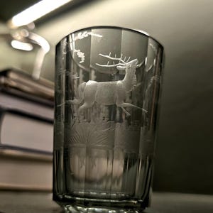 May include: A clear glass tumbler featuring an etched design of a deer in a forest setting. The glass has a slightly flared shape and a thick base. The etched design is detailed, creating a textured look. The glass is on a dark surface.