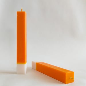 May include: Two orange and white candles with a gold wick. The candles are rectangular in shape and have a white base and an orange top.