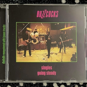 Peut inclure: Un boîtier de CD avec la pochette de l'album "Singles Going Steady" du groupe Buzzcocks. Le fond est noir avec le nom du groupe en rose et le titre de l'album en vert. L'image montre le groupe en concert.