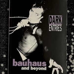 May include: A black and white photo of a man singing into a microphone. The book cover is black with white text that reads "Dark Entries Bauhaus and beyond Ian Shirley".