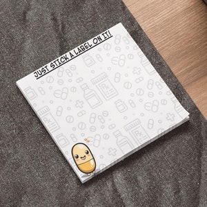 May include: A white sticky note pad with a repeating pattern of pill bottles and pills. The pad has the text "JUST STICK A LABEL ON IT!" and a cartoon pill character. The pad is on a dark gray surface.