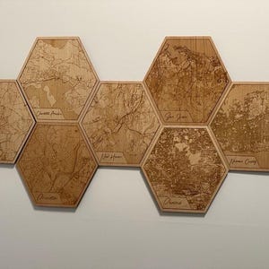 May include: A collection of seven wooden hexagon-shaped wall art pieces. Each piece features a detailed map design with city names such as "Lisle", "Charlotte Amalie", "New Haven", "San Juan", "Phoenix", and "Miami County" engraved on them. The wood has a natural, light brown color.