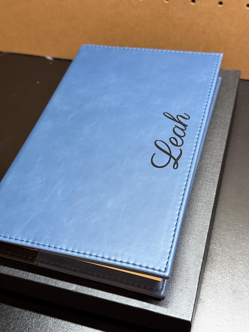 May include: A light blue leather-bound journal with the name "Leah" in cursive script. The journal is closed and rests on a dark gray box. The cover has a stitched border.