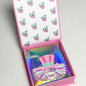 May include: A pink and iridescent box with a silver chain necklace featuring a colorful skull with a pink and white top hat and sunglasses. The box is lined with a white background with a repeating pattern of blue bears.