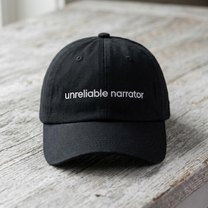Puede incluir: Gorra de béisbol negra con el texto blanco "unreliable narrator". La gorra tiene una visera curva y un botón en la parte superior. Descansa sobre una superficie de madera envejecida.