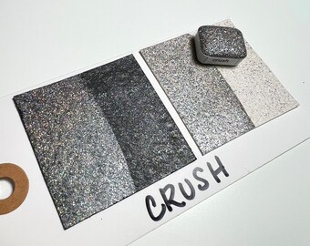 May include: A sample card with three swatches of silver glitter, labeled "CRUSH". The glitter is in different shades of silver, from light to dark. A small container of the glitter is also shown, labeled "crush".
