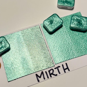 May include: A close-up of a teal green metallic paint swatch with the word "MIRTH" written in black marker. The swatch is divided into two sections, one with a lighter shade of teal green and the other with a darker shade. There are four small teal green paint pots surrounding the swatch.