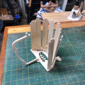 May include: A wooden, unfinished, DIY project for a baseball bat holder. The holder is made of multiple pieces of wood that are assembled together. The holder is designed to hold a baseball bat upright. The holder has the word "BEVD" engraved on the front.