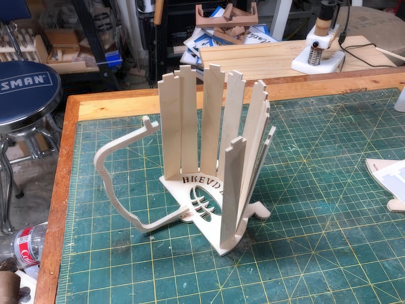 May include: A wooden, unfinished, DIY project for a baseball bat holder. The holder is made of multiple pieces of wood that are assembled together. The holder is designed to hold a baseball bat upright. The holder has the word "BEVD" engraved on the front.