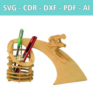May include: A light-colored wooden desk organizer featuring a cylindrical pen holder and a decorative skier figure on a curved ramp. The text SVG - CDR - DXF - PDF - AI is displayed at the top. A green pen and a red utility knife are inside the holder.