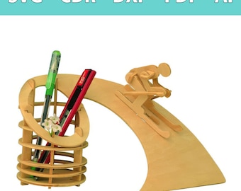 Op de afbeelding: Een bureau-organizer van lichtgekleurd hout met een cilindrische pennenhouder en een decoratief skiërsfiguur op een gebogen helling. De tekst SVG - CDR - DXF - PDF - AI staat bovenaan. Een groene pen en een rood hobbymes zitten in de houder.