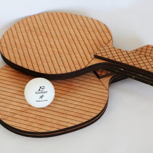 May include: Two wooden ping pong paddles with a white ball. The paddles are light brown with dark brown edges and a striped design. The ball is white with black text that reads "EastPoint" and "Made in China".
