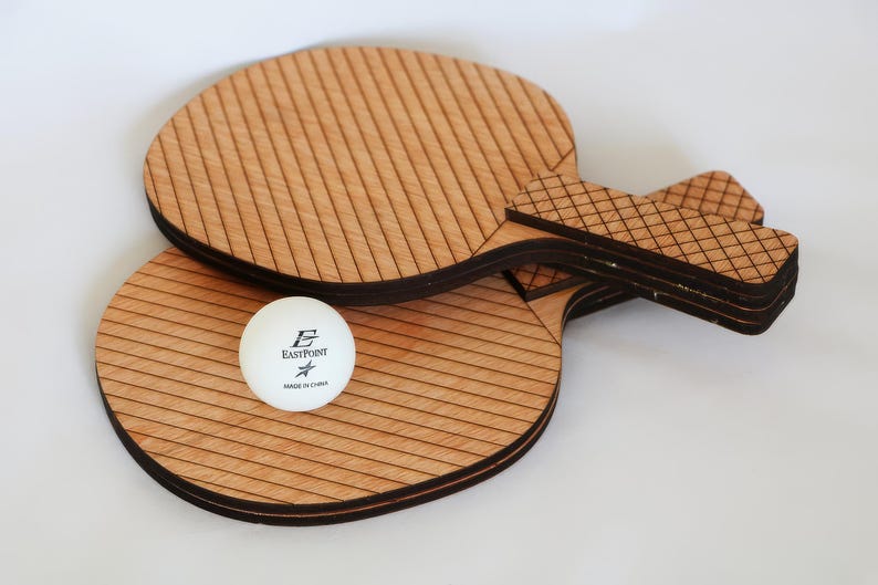 May include: Two wooden ping pong paddles with a white ball. The paddles are light brown with dark brown edges and a striped design. The ball is white with black text that reads "EastPoint" and "Made in China".
