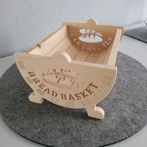 May include: A wooden bread basket with the words "BREAD BASKET" carved into the side. The basket has a cutout design of a loaf of bread and a wheat stalk.