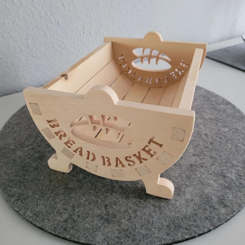 May include: A wooden bread basket with the words "BREAD BASKET" carved into the side. The basket has a cutout design of a loaf of bread and a wheat stalk.