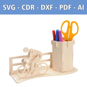 May include: A light-colored wooden desk organizer featuring a cyclist figure on a bike next to a cylindrical pen holder. The organizer includes a fence-like structure and holds three markers and yellow scissors. Text at the top reads SVG - CDR - DXF - PDF - AI.