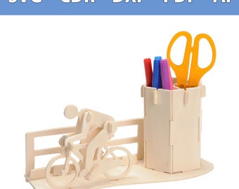 Op de afbeelding: Een bureau-organizer van lichtgekleurd hout met een fietser op een fiets naast een cilindrische pennenhouder. De organizer bevat een hekachtige structuur en bevat drie stiften en een gele schaar. De tekst bovenaan luidt SVG - CDR - DXF - PDF - AI.