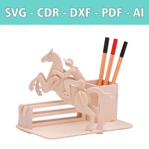 May include: A light-colored wooden desk organizer featuring a laser-cut design of a horse and rider jumping over a hurdle. The structure includes a rectangular pen holder containing three pens with black and red caps. Text reads SVG - CDR - DXF - PDF - AI.