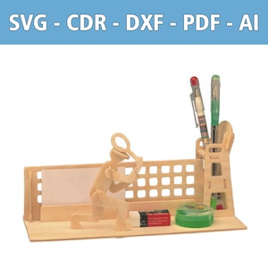 May include: A light-colored wooden desk organizer featuring a cutout figure holding a magnifying glass, a grid-patterned back panel, a pen holder containing two pens, a small rectangular eraser, and a round green container on a flat base.