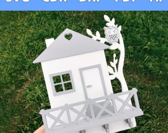 Op de afbeelding: Witte en grijze houten sleutelhanger in de vorm van een huis met een grijze uil die op een tak zit. Het huis heeft een klein raam, een deur en een grijs hek. Er zijn vijf haken aan de onderkant van het huis.