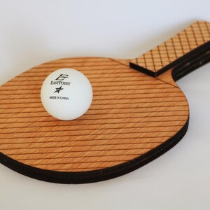May include: A wooden ping pong paddle with a white ball. The paddle has a light brown, striped surface and a dark brown handle. The white ball has the text "EASTPOINT" and "MADE IN CHINA" printed on it.