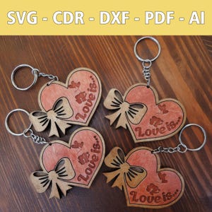 Może przedstawiać: Cztery drewniane breloki w kształcie serca z czerwonym odcieniem i tekstem "Love is..." w brązowej czcionce. Każdy brelok ma brązową kokardkę i srebrny kółko do kluczy.