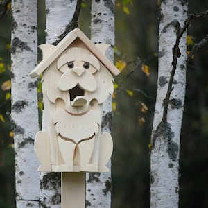 May include: A wooden birdhouse shaped like a cat with a large opening for birds to enter. The birdhouse is painted white and has black eyes and a black nose. It is mounted on a wooden pole.