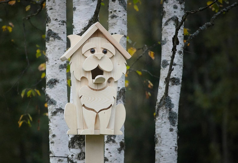 May include: A wooden birdhouse shaped like a cat with a large opening for birds to enter. The birdhouse is painted white and has black eyes and a black nose. It is mounted on a wooden pole.