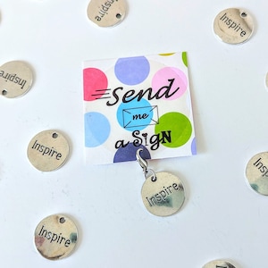 May include: A close-up of a silver charm with the word "Inspire" on it. The charm is hanging from a card that says "Send me a Sign". There are many other silver charms with the word "Inspire" on them scattered around the card.
