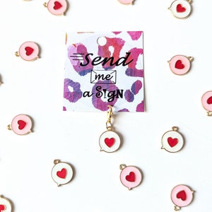May include: A white background with a variety of gold charms in the shape of a speech bubble with a red heart inside. The charms are scattered across the background. In the center of the image is a card with the text "Send me a sign" in pink and black lettering.