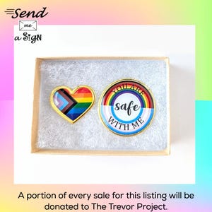 May include: Two enamel pins in a gift box. One pin is heart-shaped with a gold border and a rainbow and pride flag design. The other pin is round with a rainbow, the words "YOU ARE safe WITH ME".