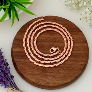 May include: A rose gold chain necklace with a textured, woven design. The necklace is displayed on a round, dark wooden surface, with a clasp at the end. The background includes sprigs of white flowers, a green plant, and a bundle of purple lavender.