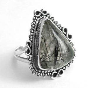 Könnte beinhalten: Ein silberner Ring mit einem dreieckigen, klaren Quarzstein mit schwarzen Einschlüssen. Der Ring hat ein detailliertes, verziertes Design um den Stein herum.