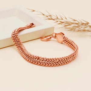 Könnte beinhalten: Ein roségoldenes Armband mit einem strukturierten, gewebten Design. Das Armband hat einen herzförmigen Verschluss. Der Schmuck wird auf einem neutralen Hintergrund präsentiert. Ein stilvolles Accessoire.