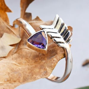 Op de afbeelding: Een zilveren ring met een amethist edelsteen in de vorm van een driehoek. De ring is versierd met een zilveren pijl met vleugels aan de zijkant.