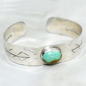 May include: A silver cuff bracelet with a turquoise stone set in the center. The bracelet is decorated with an arrow design.