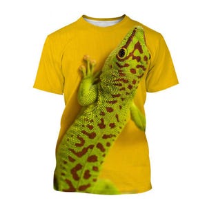 May include: Yellow t-shirt with a detailed graphic of a green and brown gecko. The gecko is positioned diagonally across the shirt. The shirt has a crew neck and short sleeves. The gecko's head and front legs are visible.
