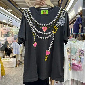 May include: A black t-shirt featuring a pearl necklace design, pink hearts, and the number 5 in yellow. The shirt has a round neckline and short sleeves. A small tag with a bear logo is visible near the neckline. Other clothing items are visible in the background.